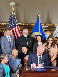 Governor Walz signs a bill surrounded by diverse people with the American and Minnesota flags in the background
