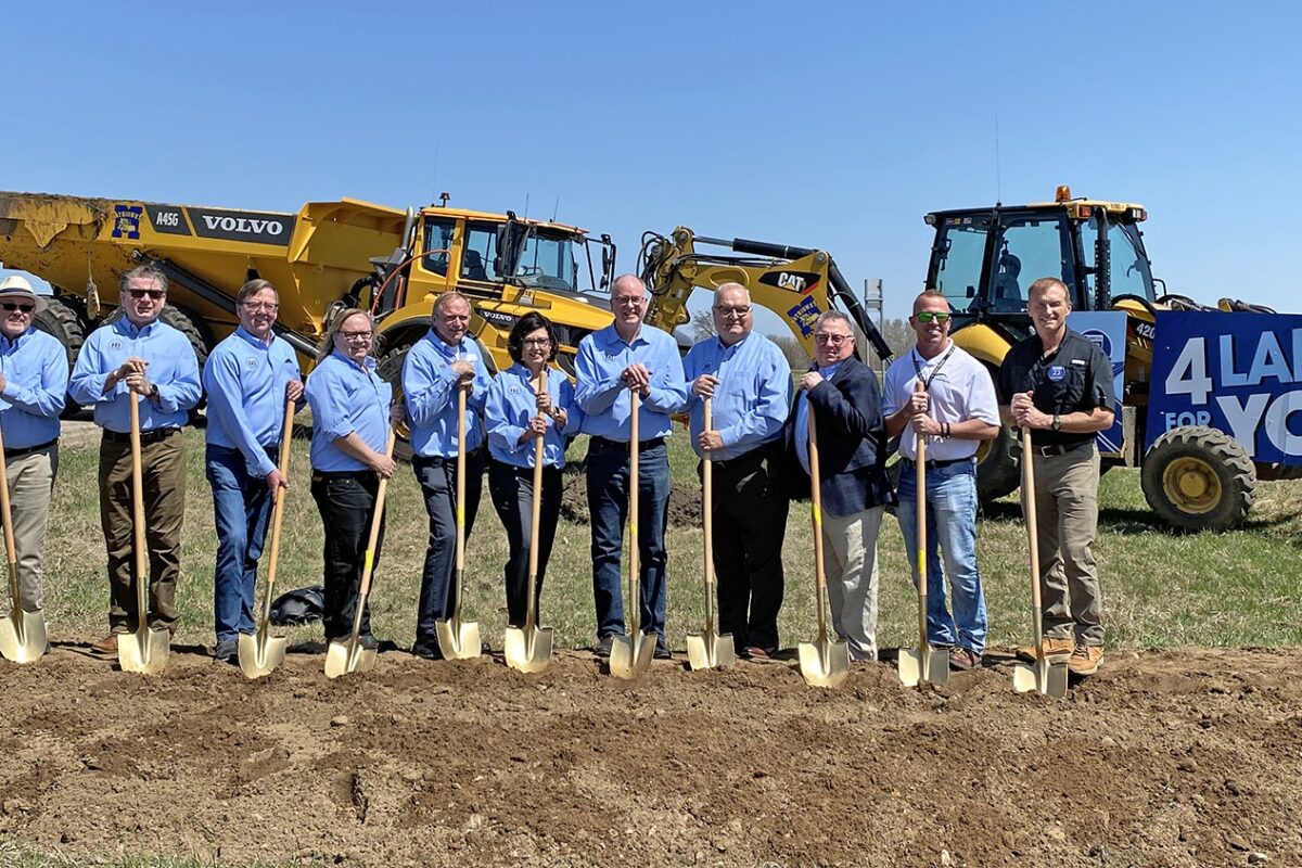 Members of the Highway 23 Highway Coalition wear matching blue polos while holding shovels. They are standing in front of a dump truck and a sign that says "4 Lanes 4 You"