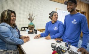 A man and woman wearing blue shirts with the 'Ag Jobs' logo stand at a counter smiling at Solange Colley